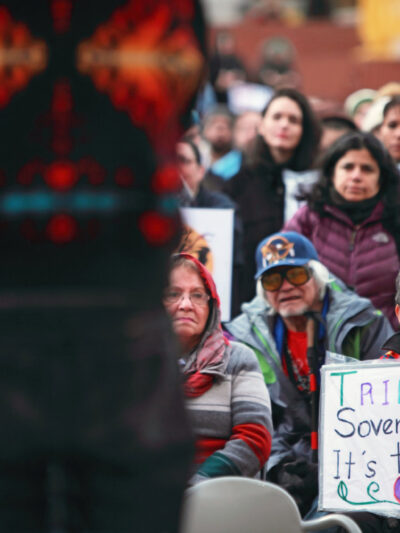 A crowd of people listens to a man speak. A woman holds a sign that reads 'Tribal Sovereignty, It's the law'