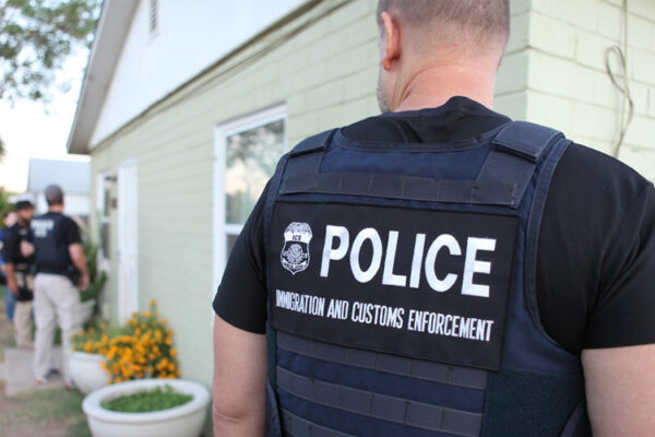 Three men wearing vests that read 'Police, Immigration and Customs Enforcement' stand in front of a house