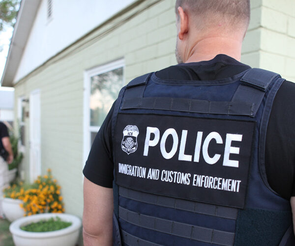Three men wearing vests that read 'Police, Immigration and Customs Enforcement' stand in front of a house
