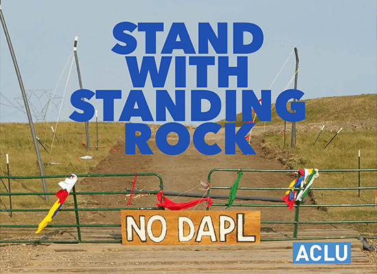 A closed metal gate stands closed with a sign reading 'No DAPL' in front of it. Text reads 'Stand with Standing Rock'