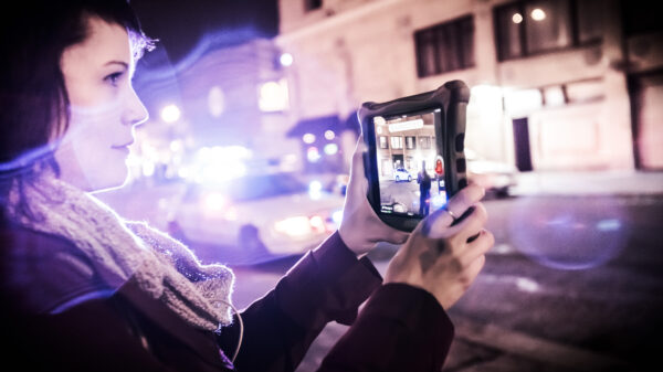 Carrie Medina holds a tablet to film the police on the street. Police lights blur into the foreground.