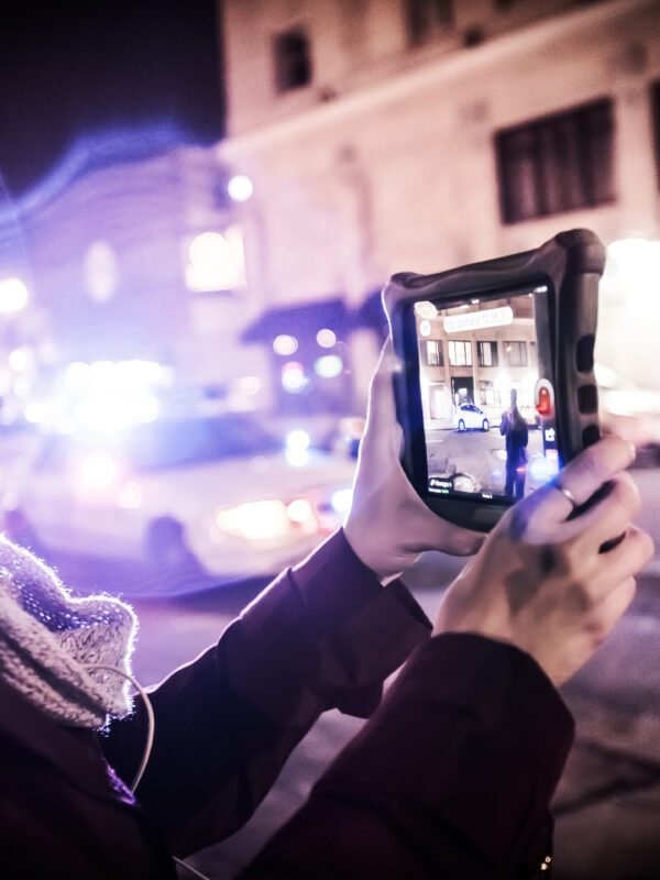 Carrie Medina holds a tablet to film the police on the street. Police lights blur into the foreground.