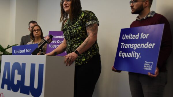 Brook Shelley stands at podium, people are holding signs in the background that have messages supporting transgender youth