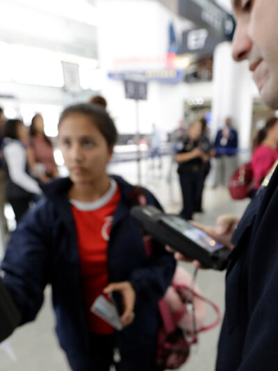 A passenger using a facial recognition kiosk in the background with a U.S. Customs and Border Protection officer watching in the foreground.