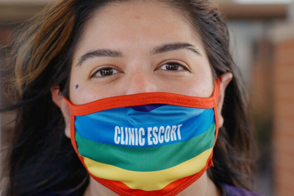 A close-up of Mariceli Alegria who's wearing a rainbow facemask with the printed words "Clinic Escort".