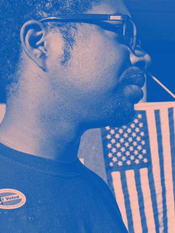 A man wears an "I Voted" stick with the American flag hanging in the background. Image tinted blue and pink.