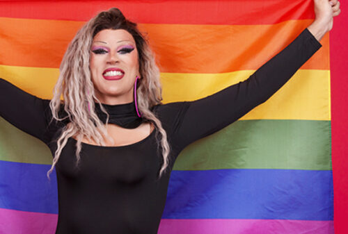 A drag queen holds up a rainbow flag with pride. Show your support by donating to the Drag Defense Fund to protect this freedom of expression.
