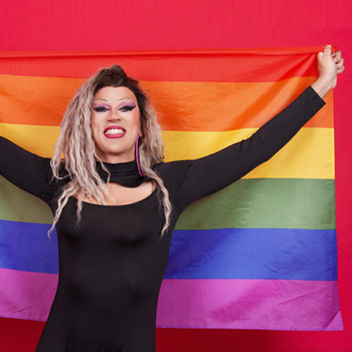 A drag queen holds up a rainbow flag with pride. Show your support by donating to the Drag Defense Fund to protect this freedom of expression.
