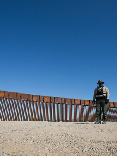 A uniformed individual in front of a portion of U.S., Mexico border wall.