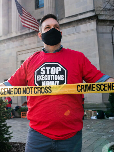 Man protesting against the death penalty in fron of the U.S. Justice Dept wearing a red shirt with a black stop sign reading "STOP EXECUTIONS NOW" in white letters.