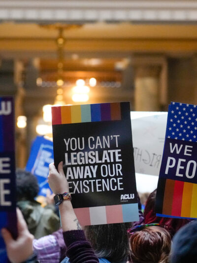 Individuals in a group holding ACLU-branded signs saying "We the People," and "You Can't Legislate Away Our Existence."