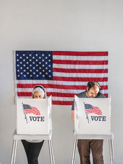 With and American flag in the background, four people in the act of voting, stand behind voter booths as they make their selections.