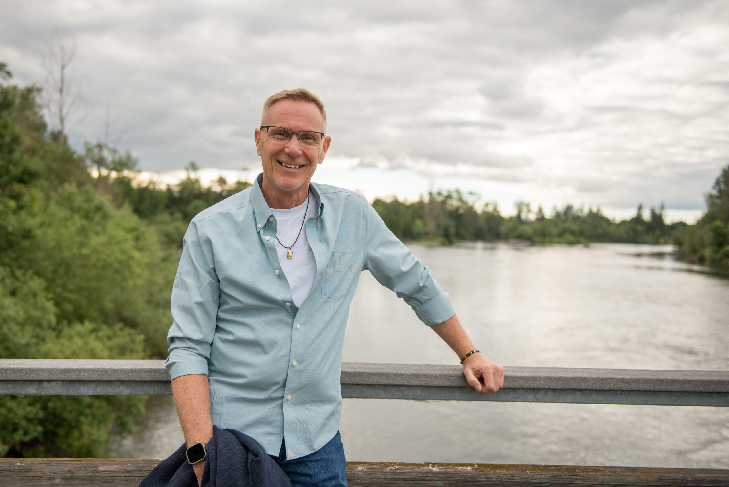 ACLU client, John Cavanaugh, a middle aged white man, smiles at the camera while standing in front of a river on a bridge. He is wearing glasses and a light blue button up shirt.
