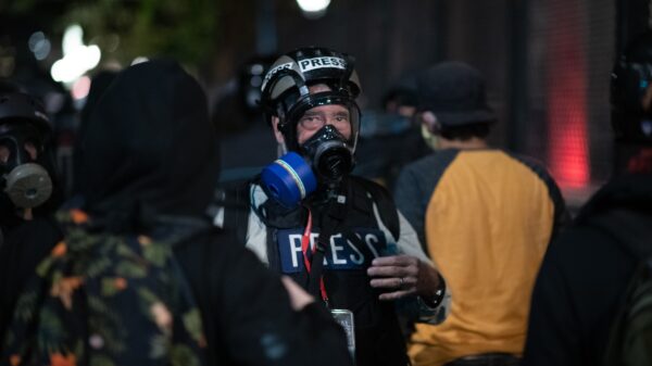 A journalist, wearing a helmet and gas mask, is standing in a crowd at a protest