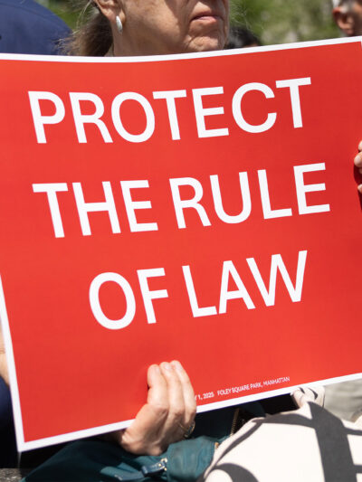 At the Lawyers Rally In Lower Manhattan To Demand Respect For The Rule Of Law, a demonstrator holds up a sign with white lettering on a red background that reds, "PROTECT THE RULE OF LAW".