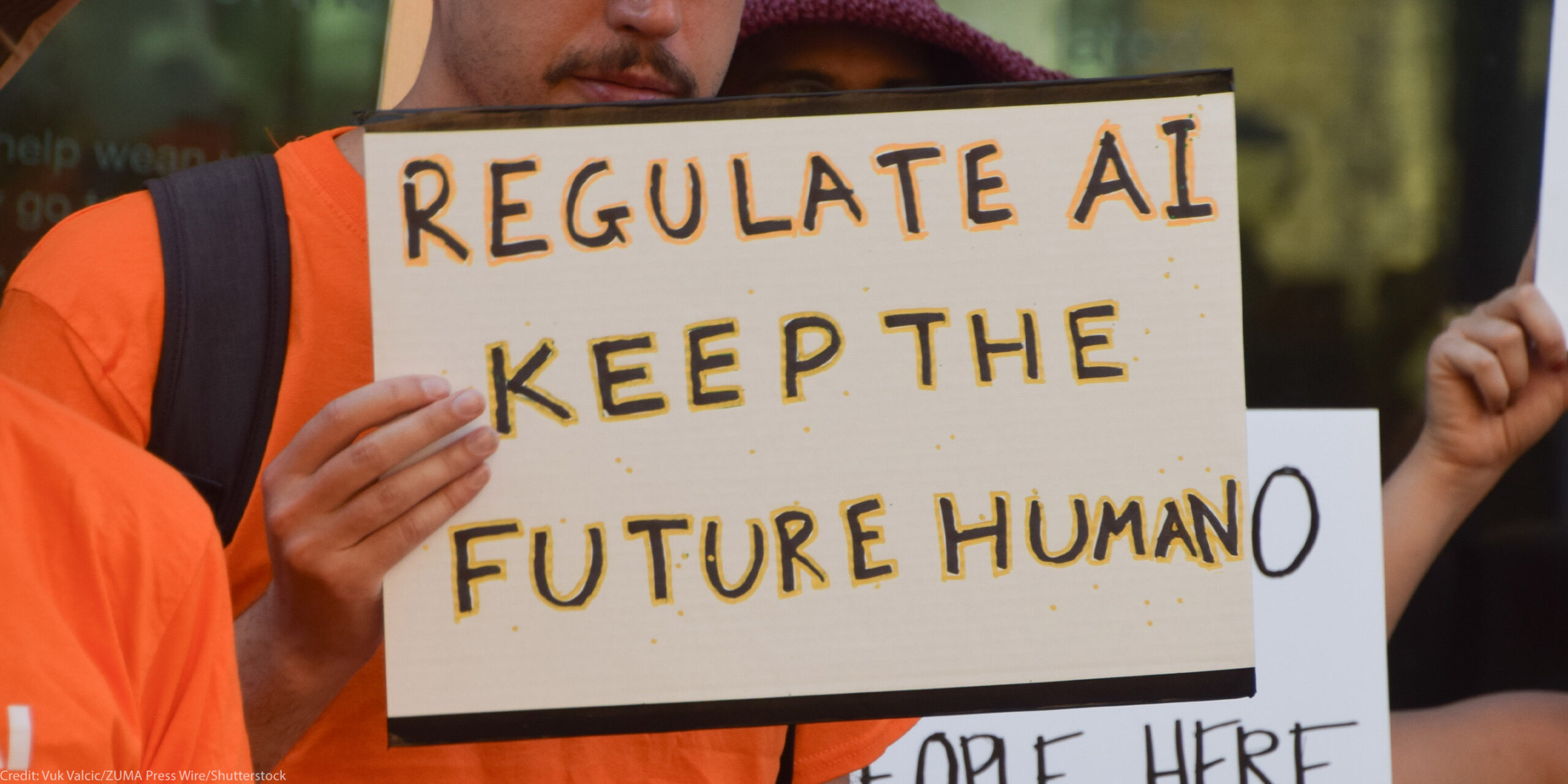 The camera focuses on a sign held by a demonstrator that reads, "REGULATE AI KEEP THE FUTURE HUMAN."