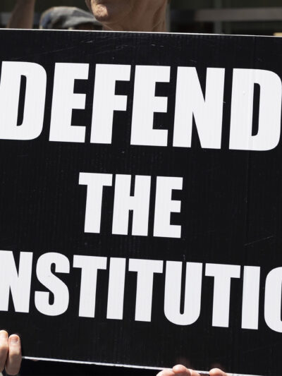 A demonstrator holds a sign (with white letters on a black background) that reads "DEFEND THE CONSTITUTION".