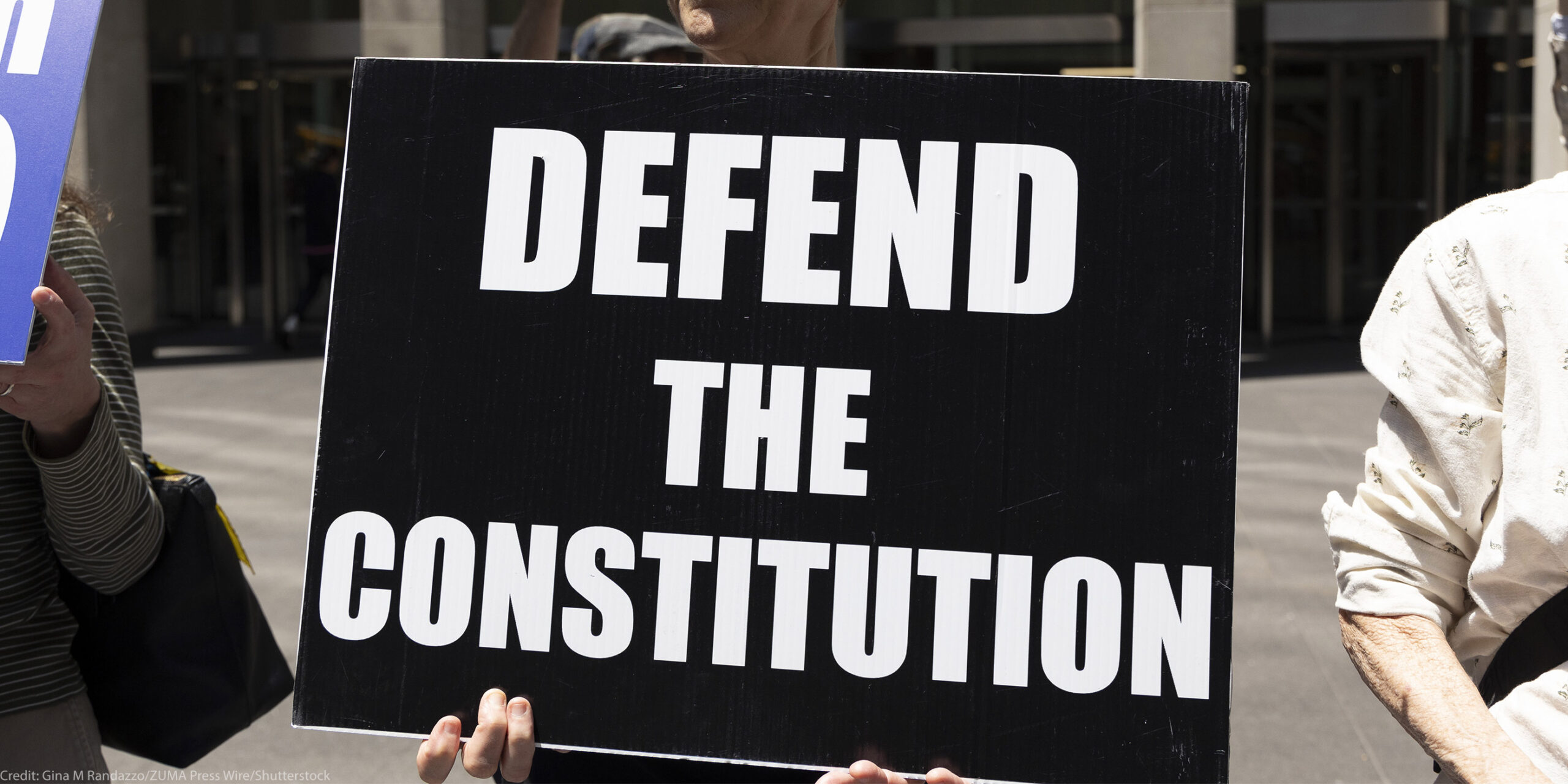 A demonstrator holds a sign (with white letters on a black background) that reads "DEFEND THE CONSTITUTION".