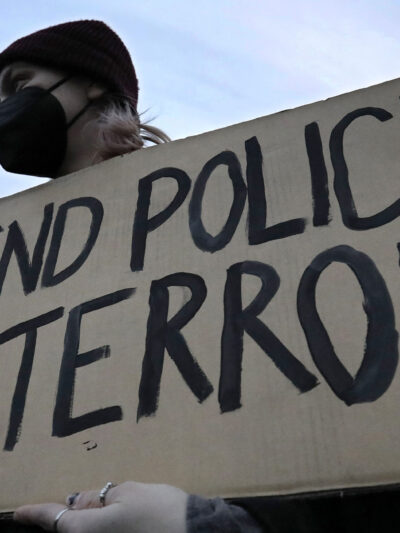 A demonstrator wearing a black hospital mask and black beanie hat holds up a cardboard sign that reads, "END POLICE TERROR".
