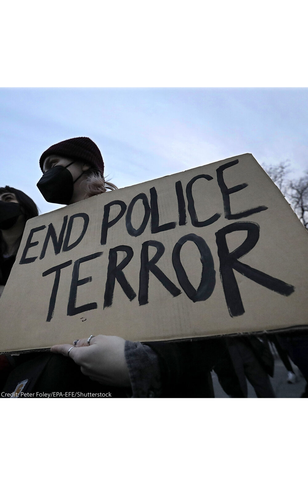 A demonstrator wearing a black hospital mask and black beanie hat holds up a cardboard sign that reads, "END POLICE TERROR".