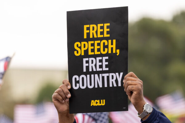 A demonstrator (with only their hands visible) holds up a sign that reads " FREE SPEECH, FREE COUNTRY - ACLU".