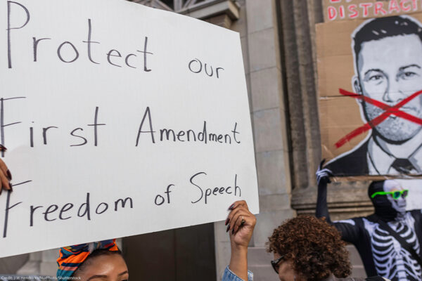 A person holds a large handwritten sign reading “Protect our First Amendment Freedom of Speech” during a protest outside the El Capitan Entertainment Centre, where Jimmy Kimmel Live! is filmed. Behind them, another protester in a skeleton costume holds a cardboard sign depicting a crossed-out image of Jimmy Kimmel. The demonstration is in response to ABC’s decision to pull Kimmel off the air following his remarks about Charlie Kirk.