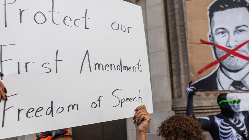 A person holds a large handwritten sign reading “Protect our First Amendment Freedom of Speech” during a protest outside the El Capitan Entertainment Centre, where Jimmy Kimmel Live! is filmed. Behind them, another protester in a skeleton costume holds a cardboard sign depicting a crossed-out image of Jimmy Kimmel. The demonstration is in response to ABC’s decision to pull Kimmel off the air following his remarks about Charlie Kirk.