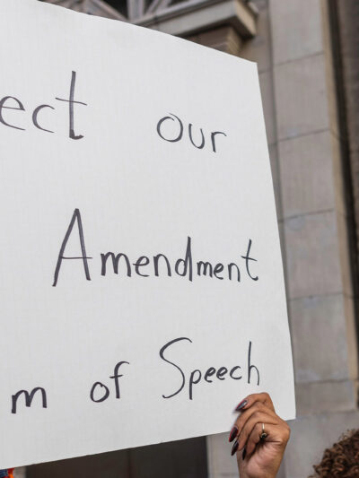 A person holds a large handwritten sign reading “Protect our First Amendment Freedom of Speech” during a protest outside the El Capitan Entertainment Centre, where Jimmy Kimmel Live! is filmed. Behind them, another protester in a skeleton costume holds a cardboard sign depicting a crossed-out image of Jimmy Kimmel. The demonstration is in response to ABC’s decision to pull Kimmel off the air following his remarks about Charlie Kirk.