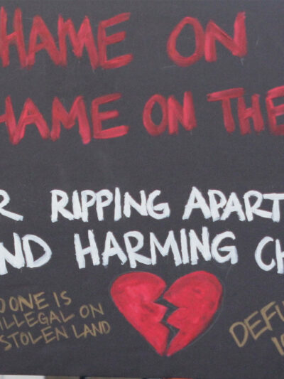 A demonstrator protests ICE and the Trump Administration policy of separating migrant families by holding up a sign (written in red ink on a black background with an illustrated broken red heart at the bottom) that reads, "SHAME ON ICE SHAME ON THE USA FOR RIPPIING APART FAMILIES AND HARMING CHILDREN No One is Illegal On Stolen Land, Defund ICE!!!"