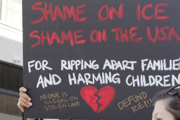 A demonstrator protests ICE and the Trump Administration policy of separating migrant families by holding up a sign (written in red ink on a black background with an illustrated broken red heart at the bottom) that reads, "SHAME ON ICE SHAME ON THE USA FOR RIPPIING APART FAMILIES AND HARMING CHILDREN No One is Illegal On Stolen Land, Defund ICE!!!"
