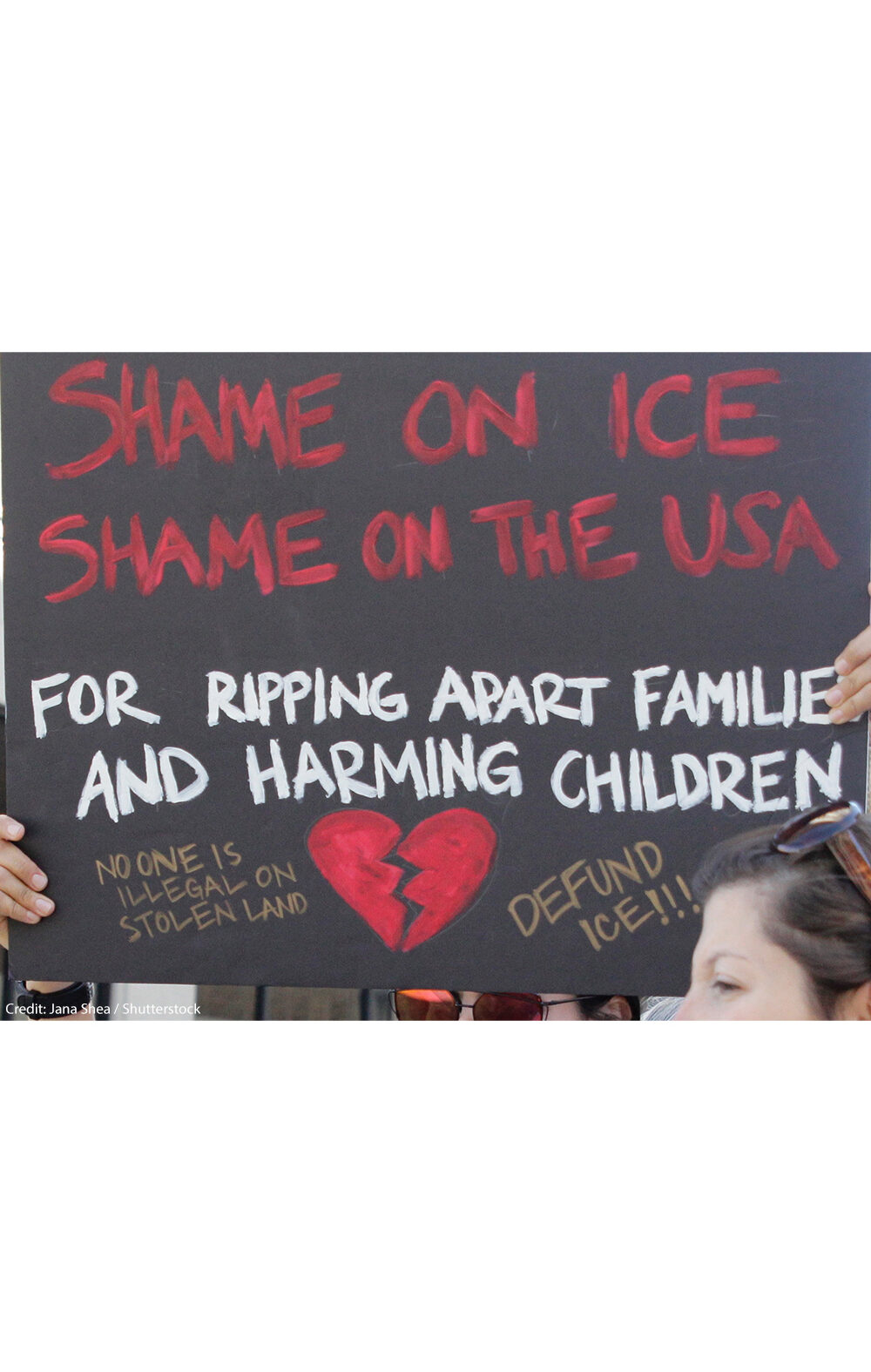 A demonstrator protests ICE and the Trump Administration policy of separating migrant families by holding up a sign (written in red ink on a black background with an illustrated broken red heart at the bottom) that reads, "SHAME ON ICE SHAME ON THE USA FOR RIPPIING APART FAMILIES AND HARMING CHILDREN No One is Illegal On Stolen Land, Defund ICE!!!"
