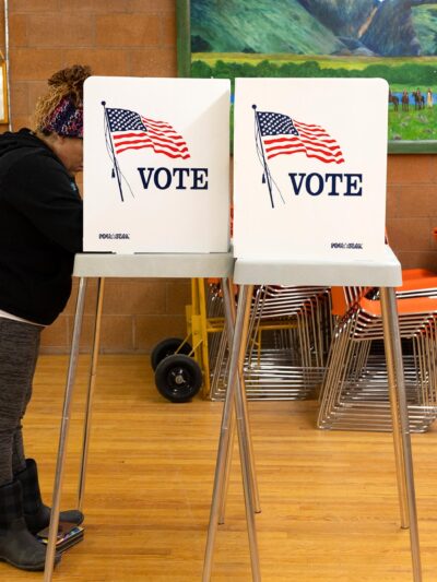 A person stands at a voting booth marked with an American flag and the word “VOTE” inside a community polling place. The room has wooden floors, orange stacked chairs, and colorful landscape paintings on the walls, with a small U.S. flag displayed nearby.