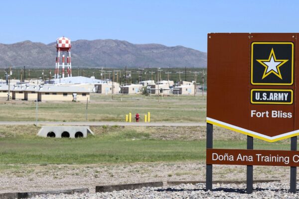 A large brown U.S. Army sign reading “Fort Bliss – Doña Ana Training Complex” stands in the foreground of a wide, open landscape. Behind it are low beige military buildings, utility poles, and a tall red-and-white checkered water tower, with mountains rising in the distance under a clear blue sky.