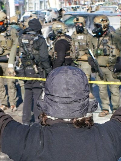 A person (with their back to the camera) in a winter hat and gloves stands with both arms raised during an anti-ICE demonstration, facing a line of police officers in tactical gear behind yellow tape on a Minneapolis street.