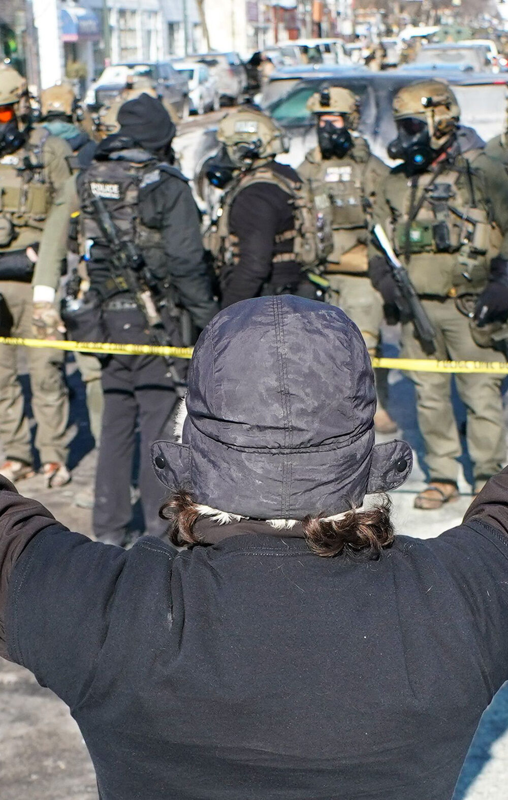 A person (with their back to the camera) in a winter hat and gloves stands with both arms raised during an anti-ICE demonstration, facing a line of police officers in tactical gear behind yellow tape on a Minneapolis street.