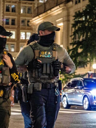 Four masked DEA police officers wearing tactical vests labeled “DEA POLICE” walk across a downtown intersection at night, with tall office buildings, streetlights, traffic signals, and cars with headlights behind them.