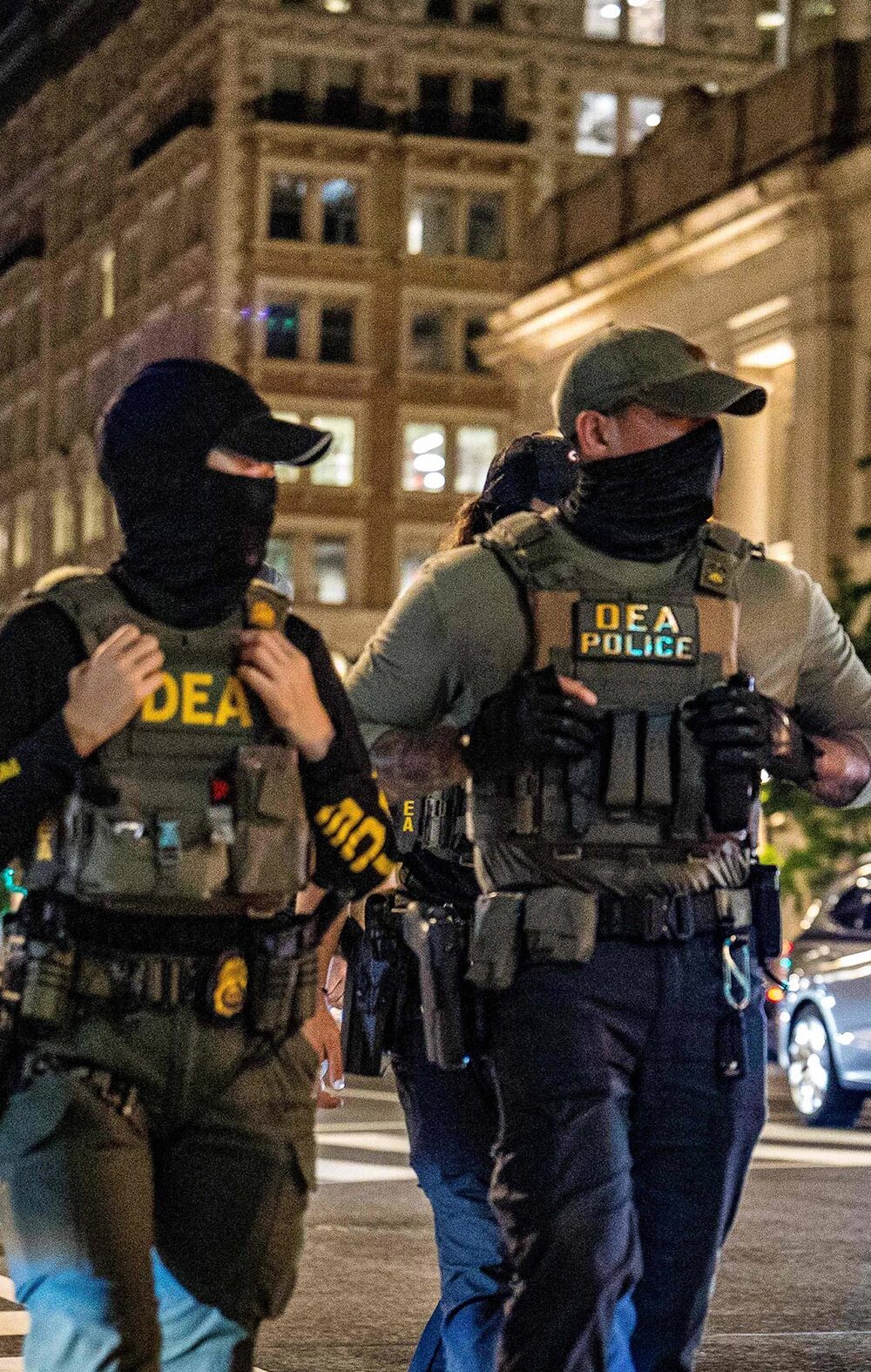 Two masked DEA police officers wearing tactical vests labeled “DEA POLICE” walk across a downtown intersection at night, with tall office buildings, streetlights, traffic signals, and cars with headlights behind them.