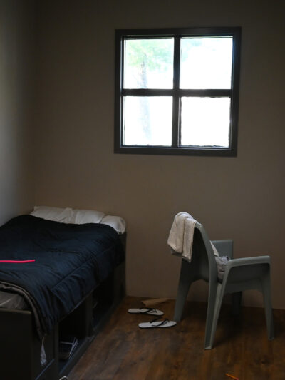 A small, sparsely furnished room with a single bed, a plastic chair with a towel draped over it, a barred window, and a pair of flip-flops on the wooden floor.
