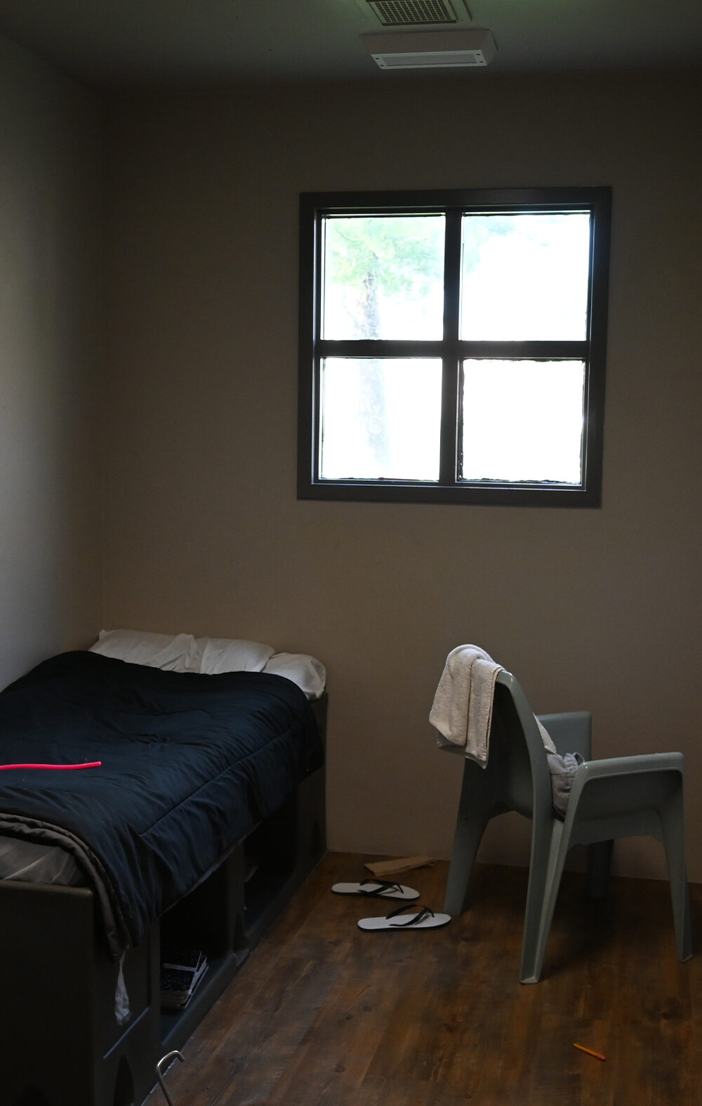 A small, sparsely furnished room with a single bed, a plastic chair with a towel draped over it, a barred window, and a pair of flip-flops on the wooden floor.