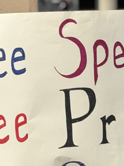 Person at a rally holds a large handmade sign reading “Free Speech, Free Press, Free People,” with a crowd and the Lincoln Memorial building in the background.