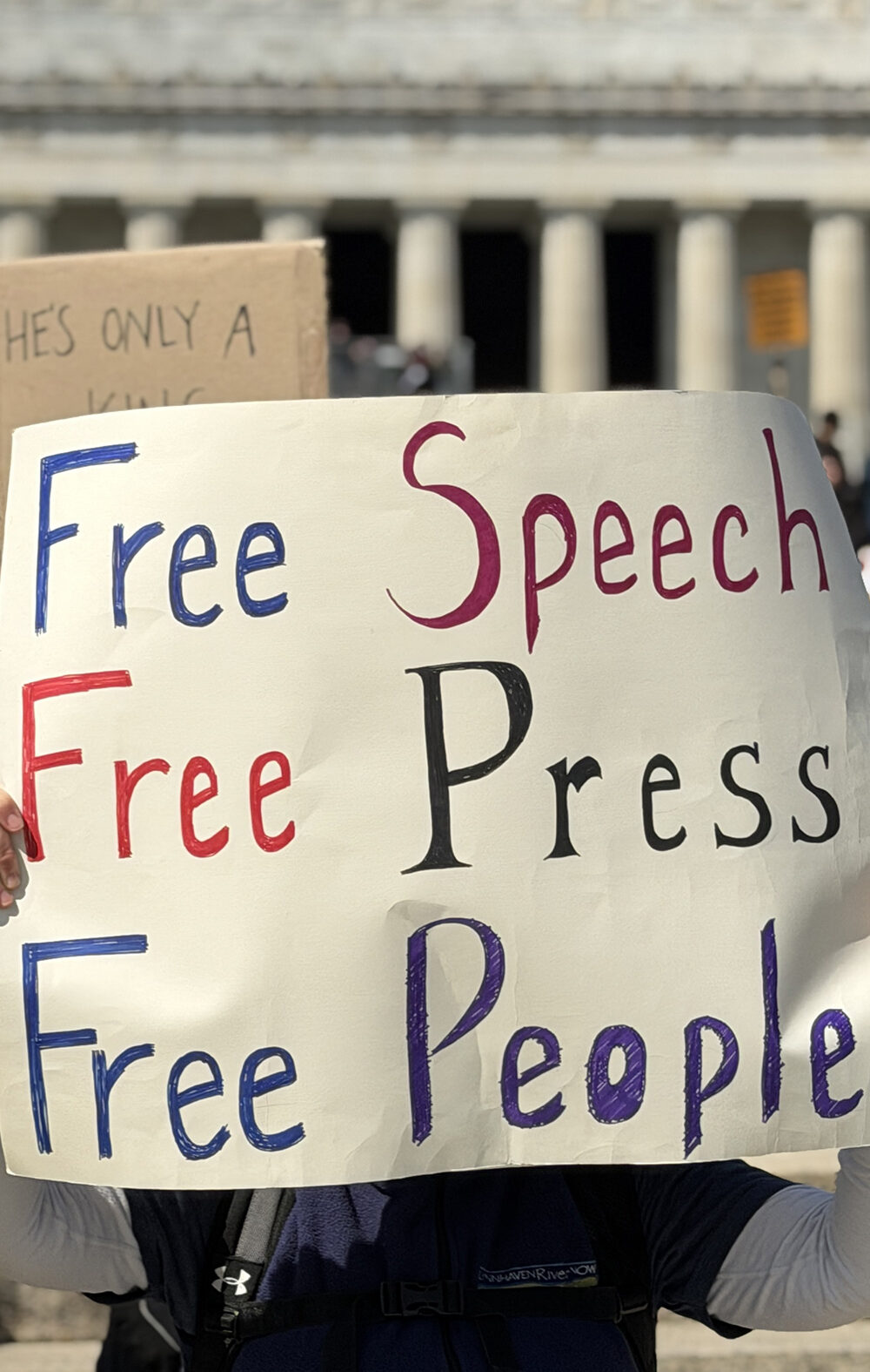 Person at a rally holds a large handmade sign reading “Free Speech, Free Press, Free People,” with a crowd and the Lincoln Memorial building in the background.