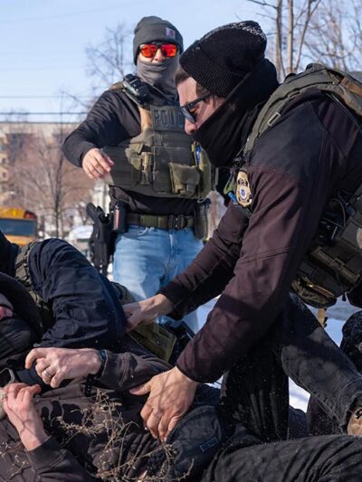 Federal immigration agents in tactical gear tackle a protester to the ground in the snow beside a chain-link fence, while other agents stand nearby in a residential area with houses and a school bus in the background.