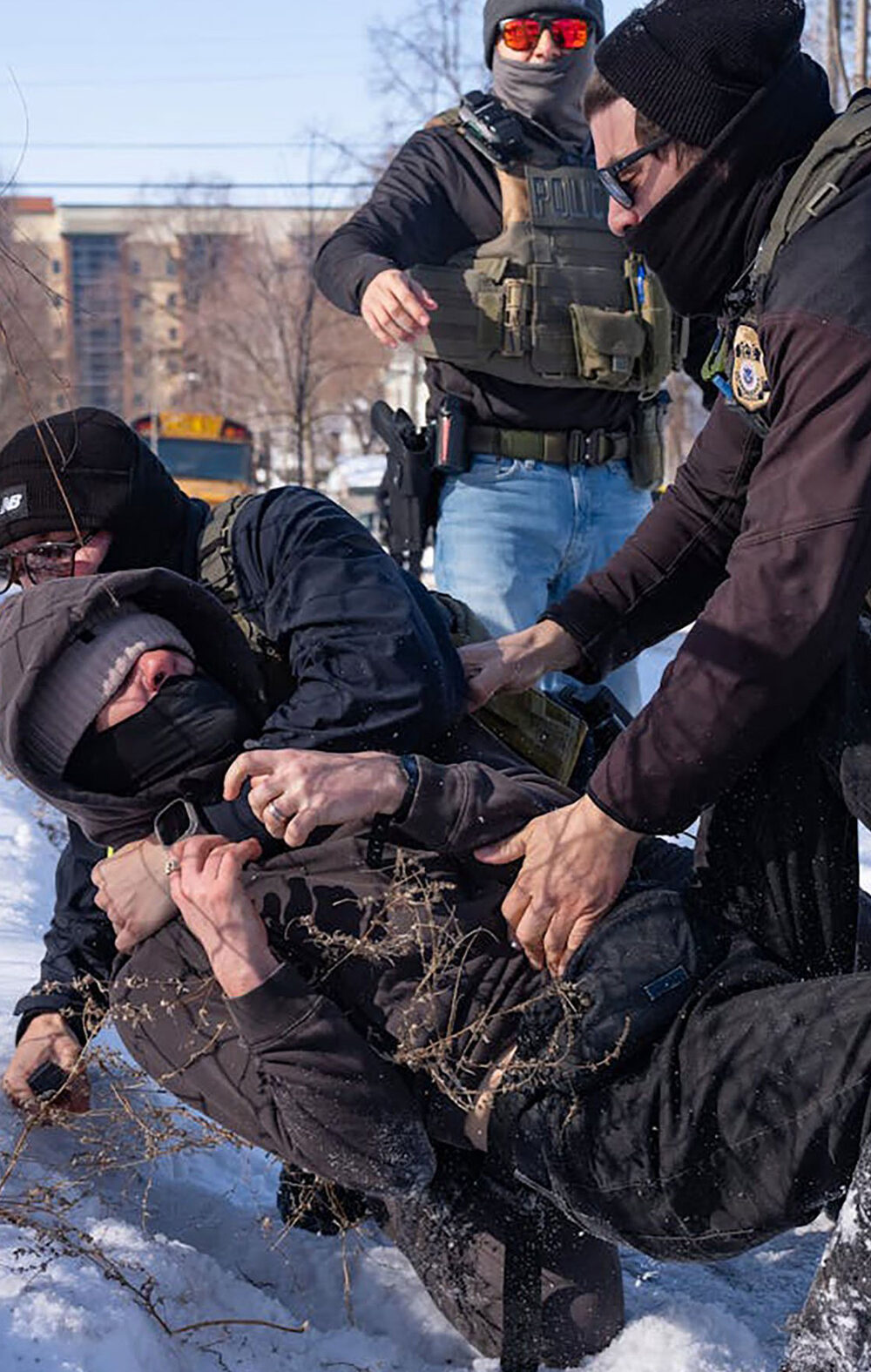 Federal immigration agents in tactical gear tackle a protester to the ground in the snow beside a chain-link fence, while other agents stand nearby in a residential area with houses and a school bus in the background.