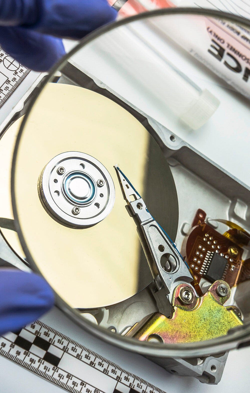 Gloved hands hold a magnifying glass over an opened computer hard drive on a forensic work surface, with evidence bags, tools, and safety glasses nearby.