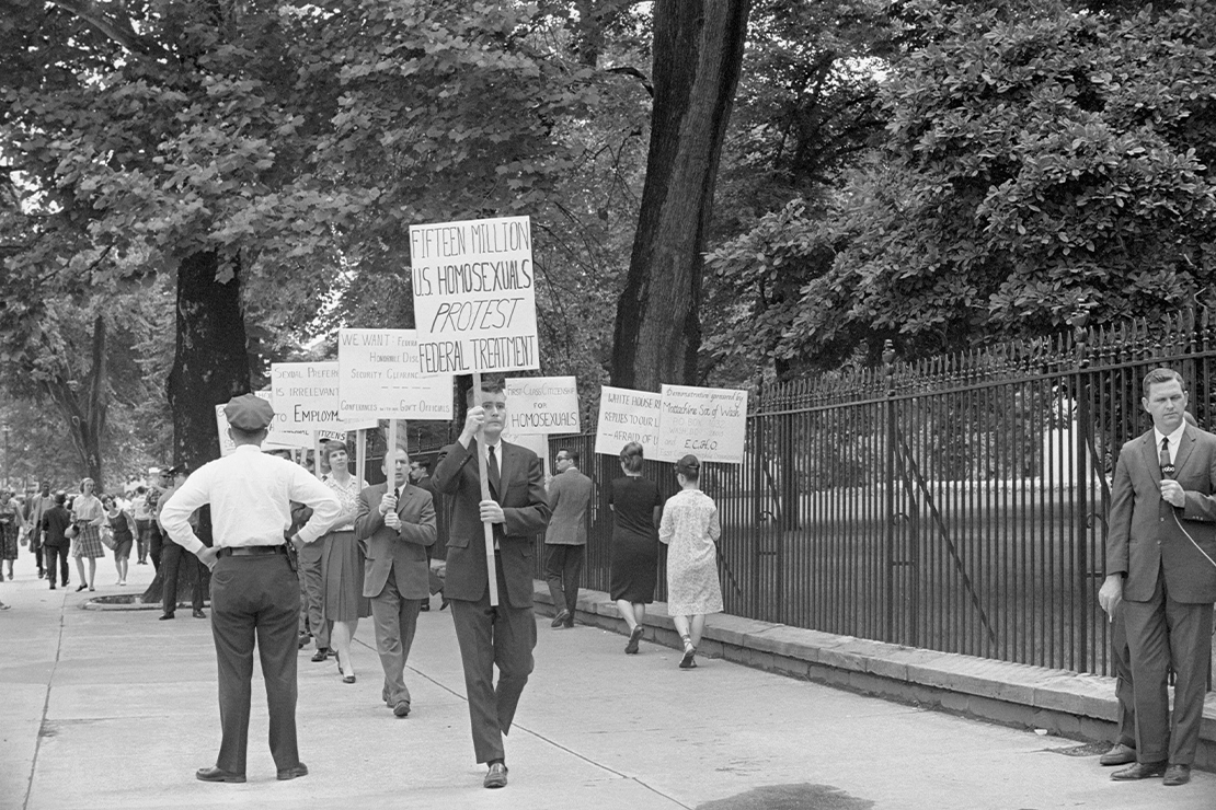 Black-and-white photo of an LGBTQ rights picket on a tree-lined sidewalk beside a tall iron fence (near the White House). Protesters (mostly in suits and dresses) carry hand-lettered signs including “FIFTEEN MILLION U.S. HOMOSEXUALS PROTEST FEDERAL TREATMENT,” “FIRST-CLASS CITIZENSHIP FOR HOMOSEXUALS,” and “SEXUAL PREFERENCE IS IRRELEVANT TO EMPLOYMENT.” A uniformed police officer stands with hands on hips facing the group, while an ABC TV reporter in a suit holds a microphone at the curb; pedestrians walk past in the background.