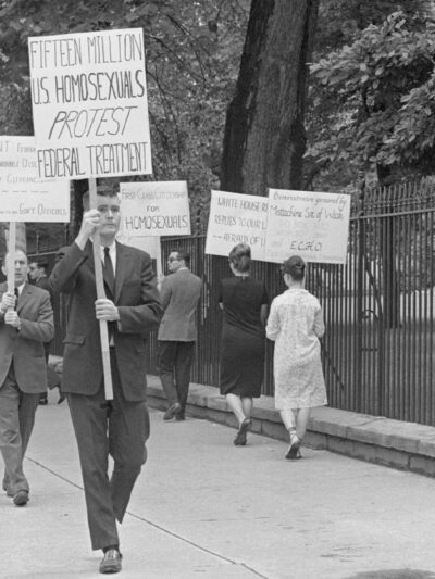 Black-and-white photo of an LGBTQ rights picket on a tree-lined sidewalk beside a tall iron fence (near the White House). Protesters—mostly in suits and dresses—carry hand-lettered signs including “FIFTEEN MILLION U.S. HOMOSEXUALS PROTEST FEDERAL TREATMENT,” “FIRST-CLASS CITIZENSHIP FOR HOMOSEXUALS,” and “SEXUAL PREFERENCE IS IRRELEVANT TO EMPLOYMENT.” A uniformed police officer stands with hands on hips facing the group, while an ABC TV reporter in a suit holds a microphone at the curb; pedestrians walk past in the background.