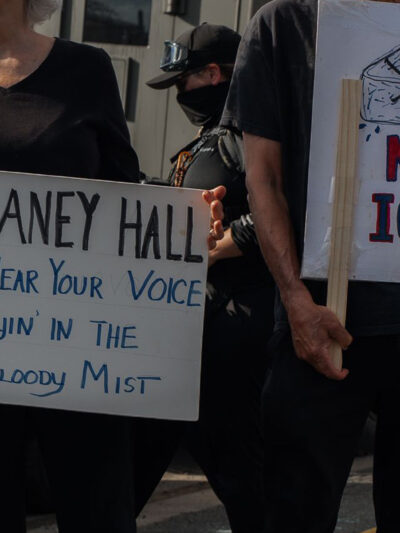 People hold signs outside the Delaney Hall Detention Facility during a protest against the treatment of detainees; one sign reads "Delaney Hall, we hear your voice cryin’ in the bloody mist," and another says "NO ICE" beneath a skull-and-crossbones drawing, with a masked officer and parked vehicles behind them.