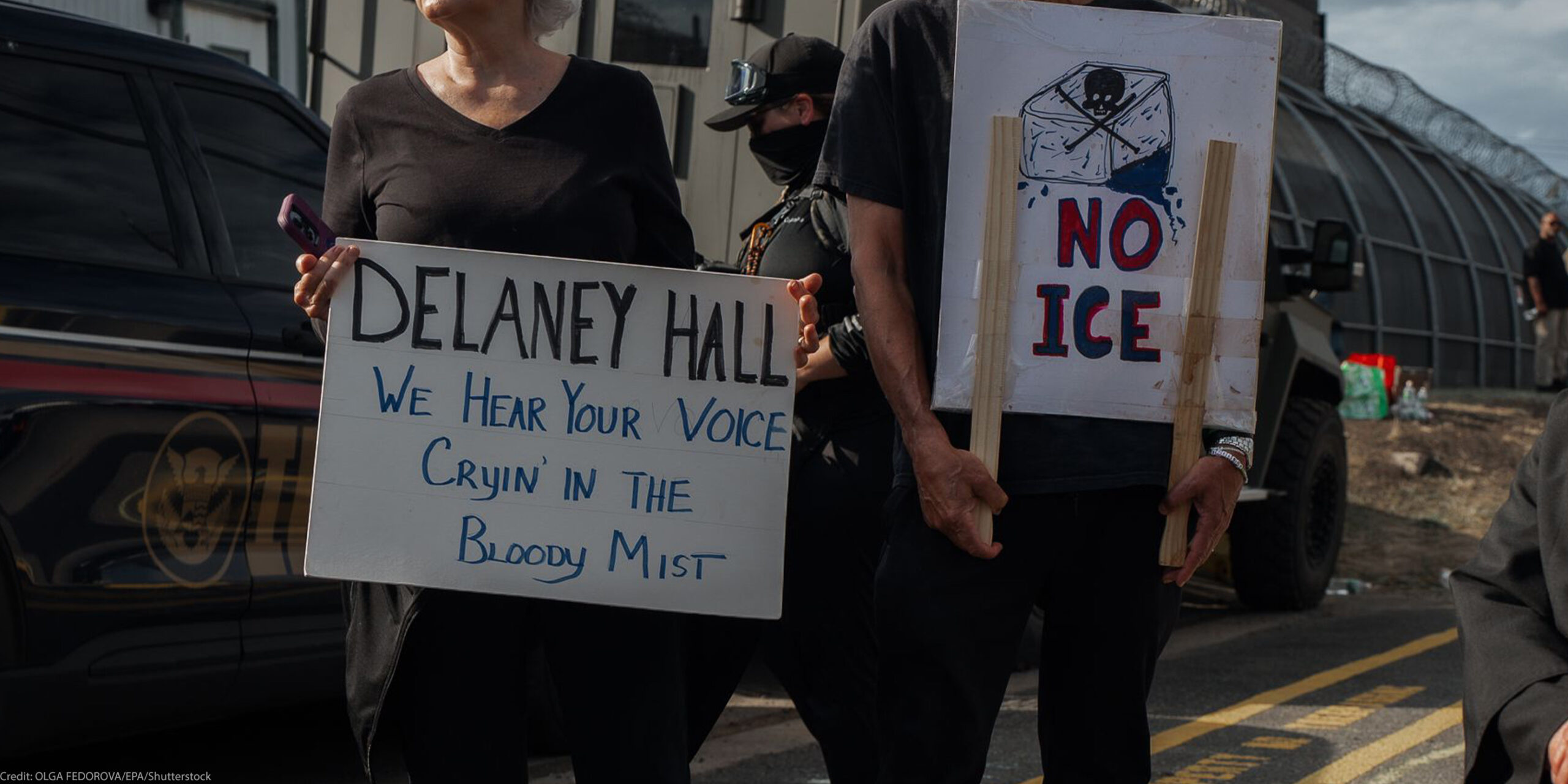 People hold signs outside the Delaney Hall Detention Facility during a protest against the treatment of detainees; one sign reads "Delaney Hall, we hear your voice cryin’ in the bloody mist," and another says "NO ICE" beneath a skull-and-crossbones drawing, with a masked officer and parked vehicles behind them.