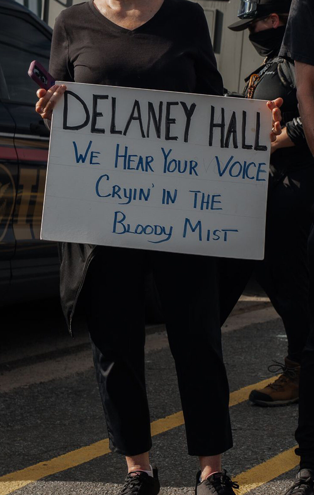 People hold signs outside the Delaney Hall Detention Facility during a protest against the treatment of detainees; one sign reads "Delaney Hall, we hear your voice cryin’ in the bloody mist," and another says "NO ICE" beneath a skull-and-crossbones drawing, with a masked officer and parked vehicles behind them.