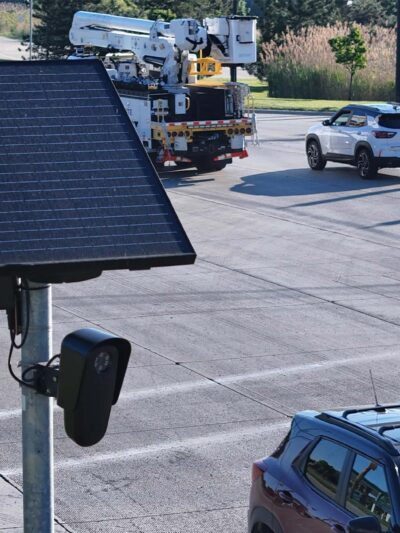A solar-powered Flock camera is mounted on a pole beside a busy multi-lane road, with cars and a utility truck driving through the intersection in the background.